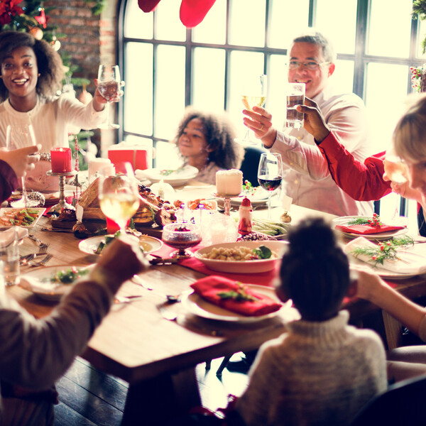A photo of a multiracial and multi-generational family together at a dinner table. The adults are holding up their glasses toward the center. There is a Christmas tree and stockings in the background.