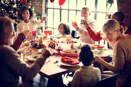 A photo of a multiracial and multi-generational family together at a dinner table. The adults are holding up their glasses toward the center. There is a Christmas tree and stockings in the background.