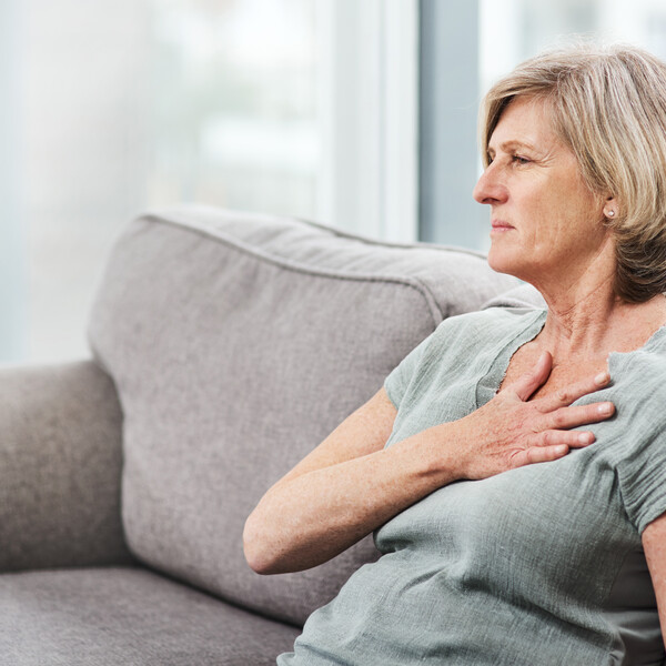 A white woman with short gray hair sits on a couch with her hand on her chest as though having chest pains