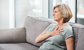 A white woman with short gray hair sits on a couch with her hand on her chest as though having chest pains
