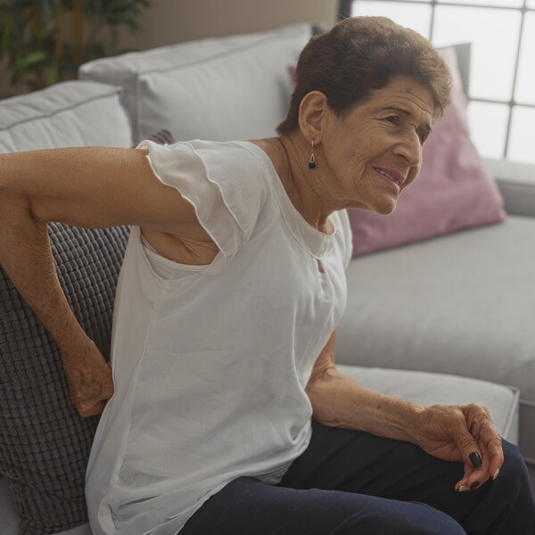 An older Latina woman sits on a couch with her had on her back as though experiencing back pain. She has short hair and is wearing a white t-shirt and dark pants.