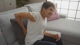 An older Latina woman sits on a couch with her had on her back as though experiencing back pain. She has short hair and is wearing a white t-shirt and dark pants.