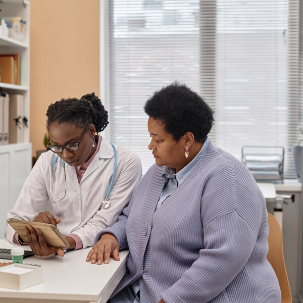 A Black woman doctor sits with her patient, reviewing medical results on a tablet
