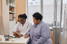 A Black woman doctor sits with her patient, reviewing medical results on a tablet