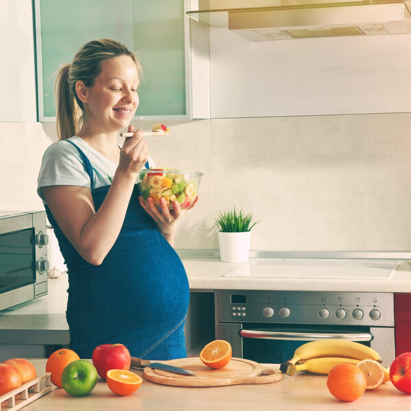 A pregnant woman stands in a kitchen eating a bowl of fruit