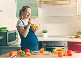 A pregnant woman stands in a kitchen eating a bowl of fruit