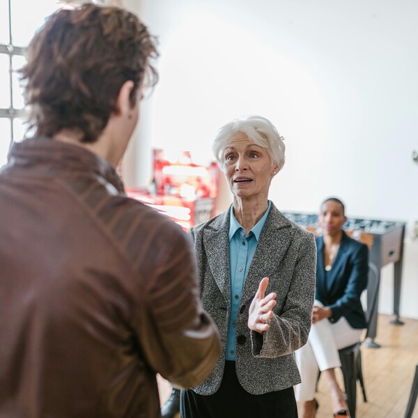 The back of a younger white man in a brown leather jacket speaking with an older white woman in a blazer