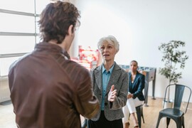 The back of a younger white man in a brown leather jacket speaking with an older white woman in a blazer
