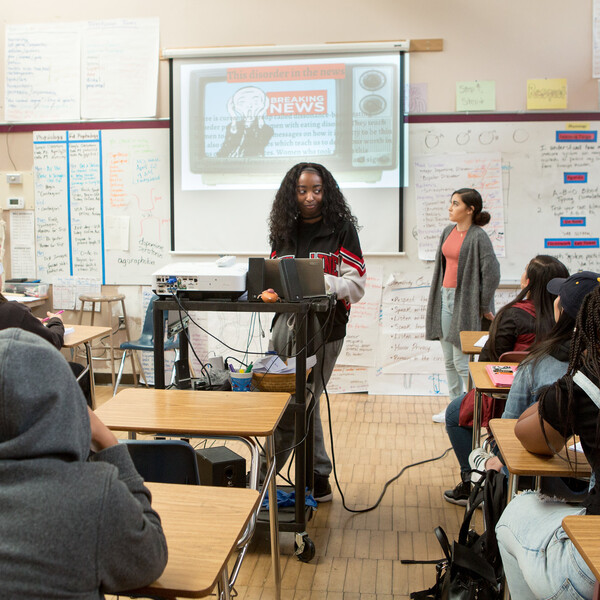A Black girl stands in front of her class as though presenting to the other students.