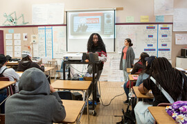 A Black girl stands in front of her class as though presenting to the other students.