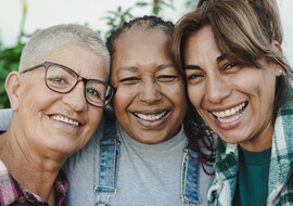 The faces of three older women of varying racial/ethnicities standing together and smiling.