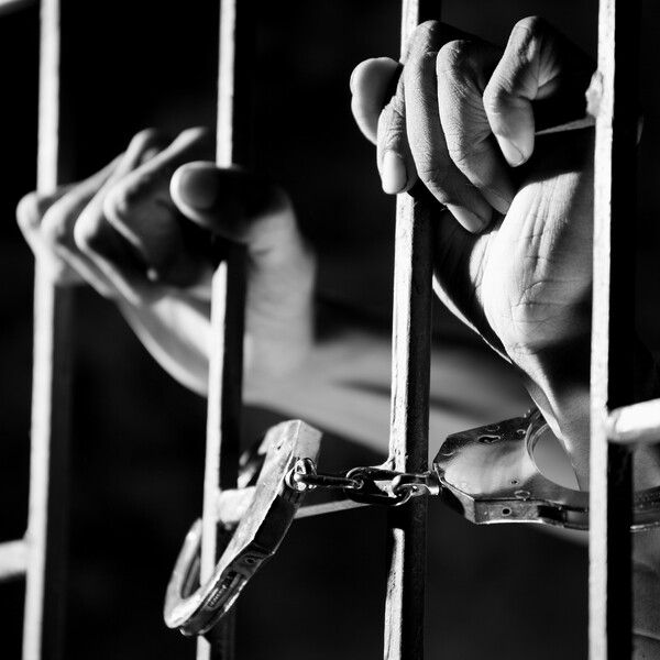 A black and white photo of handcuffed hands holding prison bars