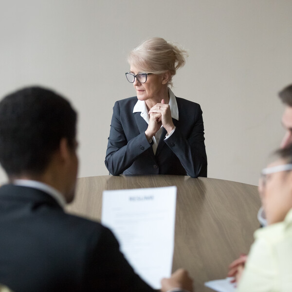 An older woman sits alone at the end of a table opposite a group of four people looking at a sheet of paper
