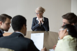 An older woman sits alone at the end of a table opposite a group of four people looking at a sheet of paper