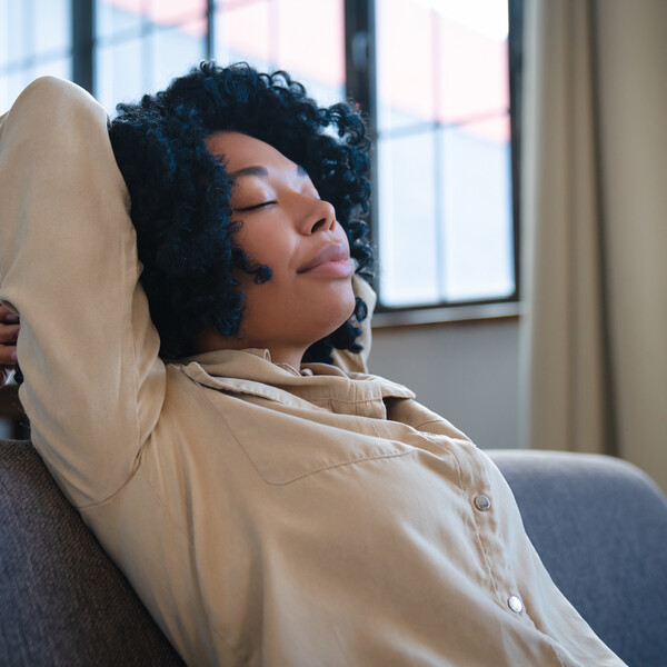 A Black woman sits on a couch resting with her hands behind her head and her eyes closed