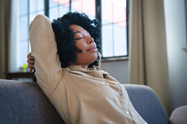 A Black woman sits on a couch resting with her hands behind her head and her eyes closed