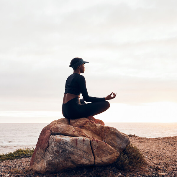 A Black woman meditates on a rock looking out on a lake