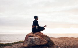 A Black woman meditates on a rock looking out on a lake