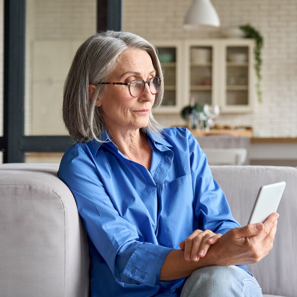 An older white woman sits on her couch looking at her phone