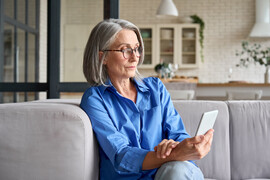An older white woman sits on her couch looking at her phone