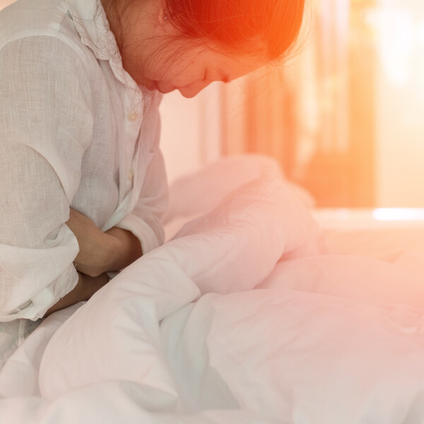 A woman sits on a bed, clasping her midsection as though in pain