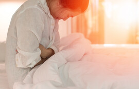 A woman sits on a bed, clasping her midsection as though in pain