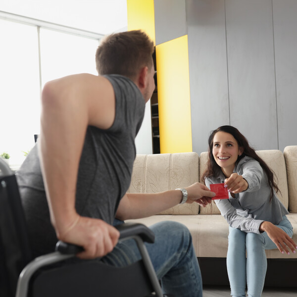 A woman on a couch hands a condom to a man in a wheelchair in front of her.