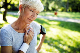 A close up of an older woman standing outside holds her hand over her chest