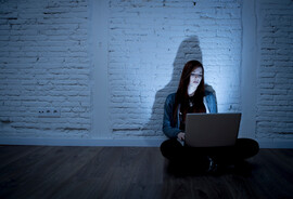 A woman sits on a floor looking at an open laptop that's in her lap
