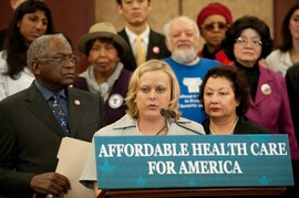 A woman stands at a podium speaking with a crowd of people behind her. The blue sign on the podium reads the following text in white: "Affordable Health Care for America."