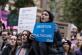 A crowd of people with a woman who appears to be on someone's shoulders holding a blue sign that reads: "Abortion is Healthcare"