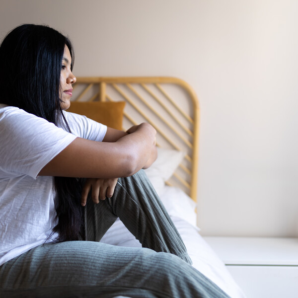 A woman who appears sad sits on a bed in pajamas