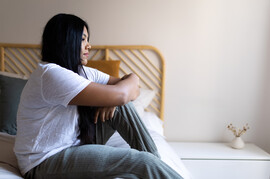 A woman who appears sad sits on a bed in pajamas