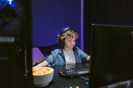 A white boy with curly hair sits on a computer in a dark room