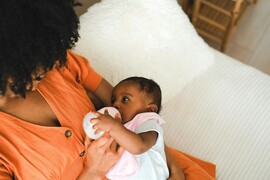 A Black woman with curly hair feeds a baby with a bottle