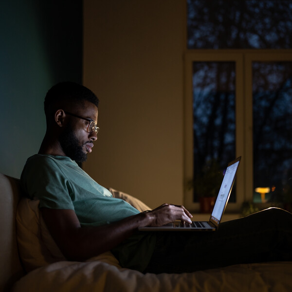 A Black man in glasses sits on a bed with a laptop on his lap