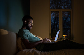A Black man in glasses sits on a bed with a laptop on his lap