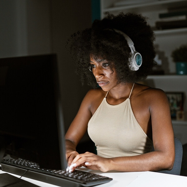 A Black woman wearing headphones sits on a computer in a dark room