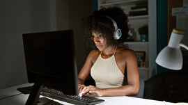 A Black woman wearing headphones sits on a computer in a dark room