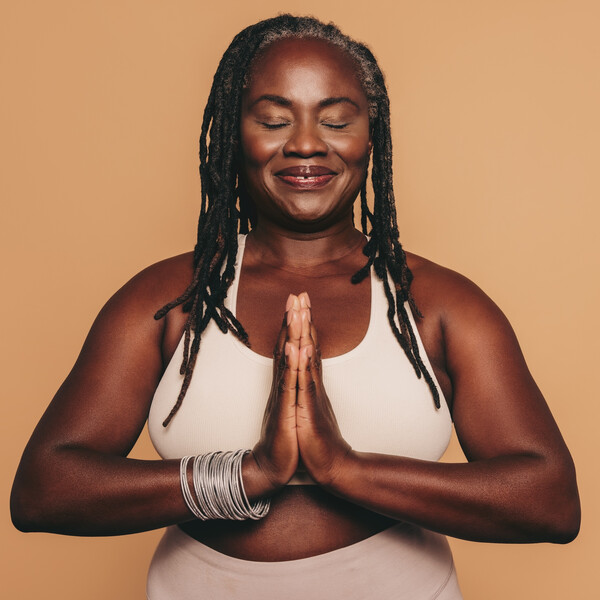 Mature woman meditating with her eyes closed and her hands in prayer position. Black woman with dreadlocks practicing yoga in a studio. Happy middle-aged woman maintaining a healthy lifestyle.