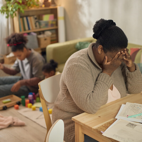 African American mother covering face while sitting by unpaid bills, children playing behind her at home.