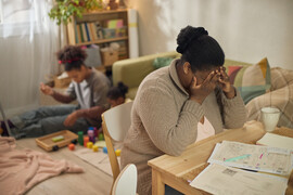 African American mother covering face while sitting by unpaid bills, children playing behind her at home.