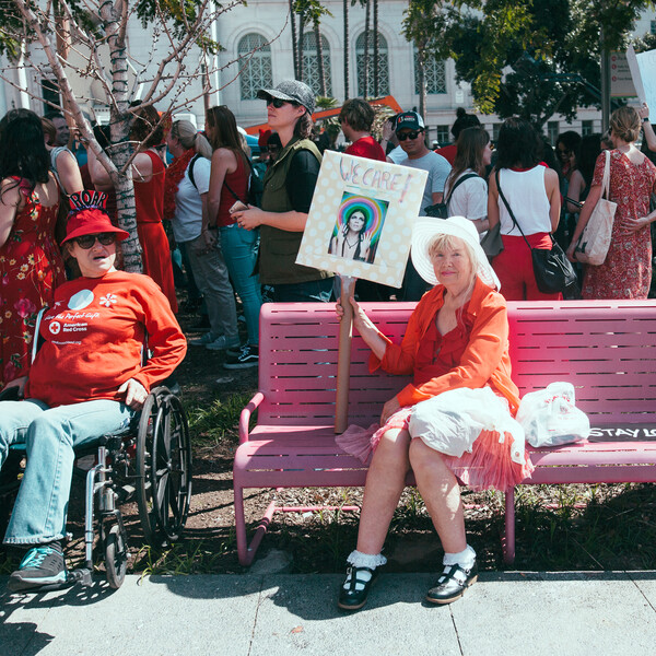 Protestors assembled, with two wheelchair users in the foreground with a woman seated on a pink hat holding up a sign reading "We Care."
