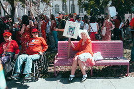Protestors assembled, with two wheelchair users in the foreground with a woman seated on a pink hat holding up a sign reading "We Care."