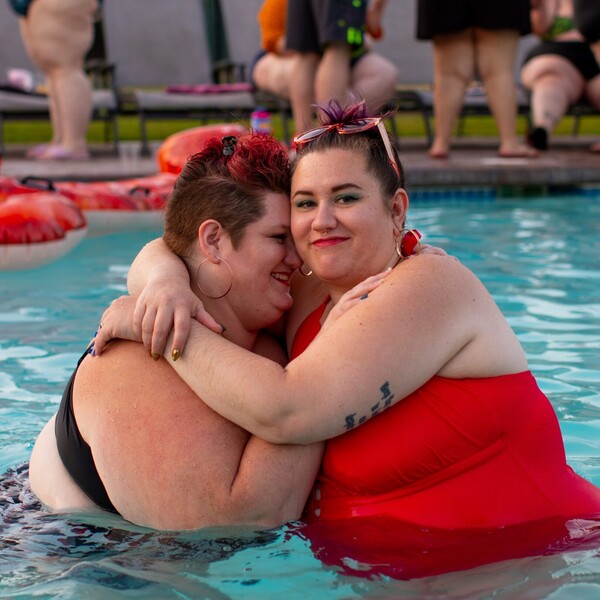 Two plus size women in a pool. One is wearing a black bikini and hugging the other who is wearing a red one piece swimsuit.