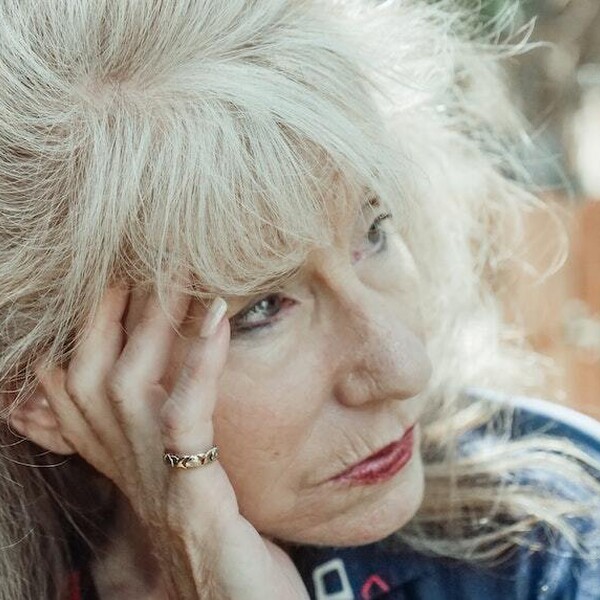 The face of an older woman with white hair and bangs. She looks pensive. She is sitting outside. She is wearing red lipstick and a blue sweatshirt.
