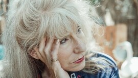 The face of an older woman with white hair and bangs. She looks pensive. She is sitting outside. She is wearing red lipstick and a blue sweatshirt.