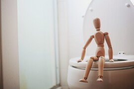 A wooden toy person sits on a white toilet.