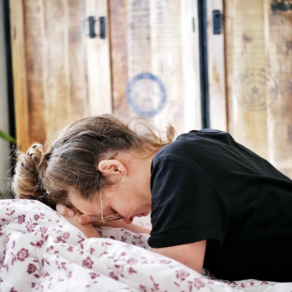 A woman is on her bed, she leans forward with her head in her hands.
