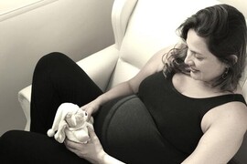 A black and white photo, a pregnant woman wearing a black top and black leggings is holding a small teddy bear.